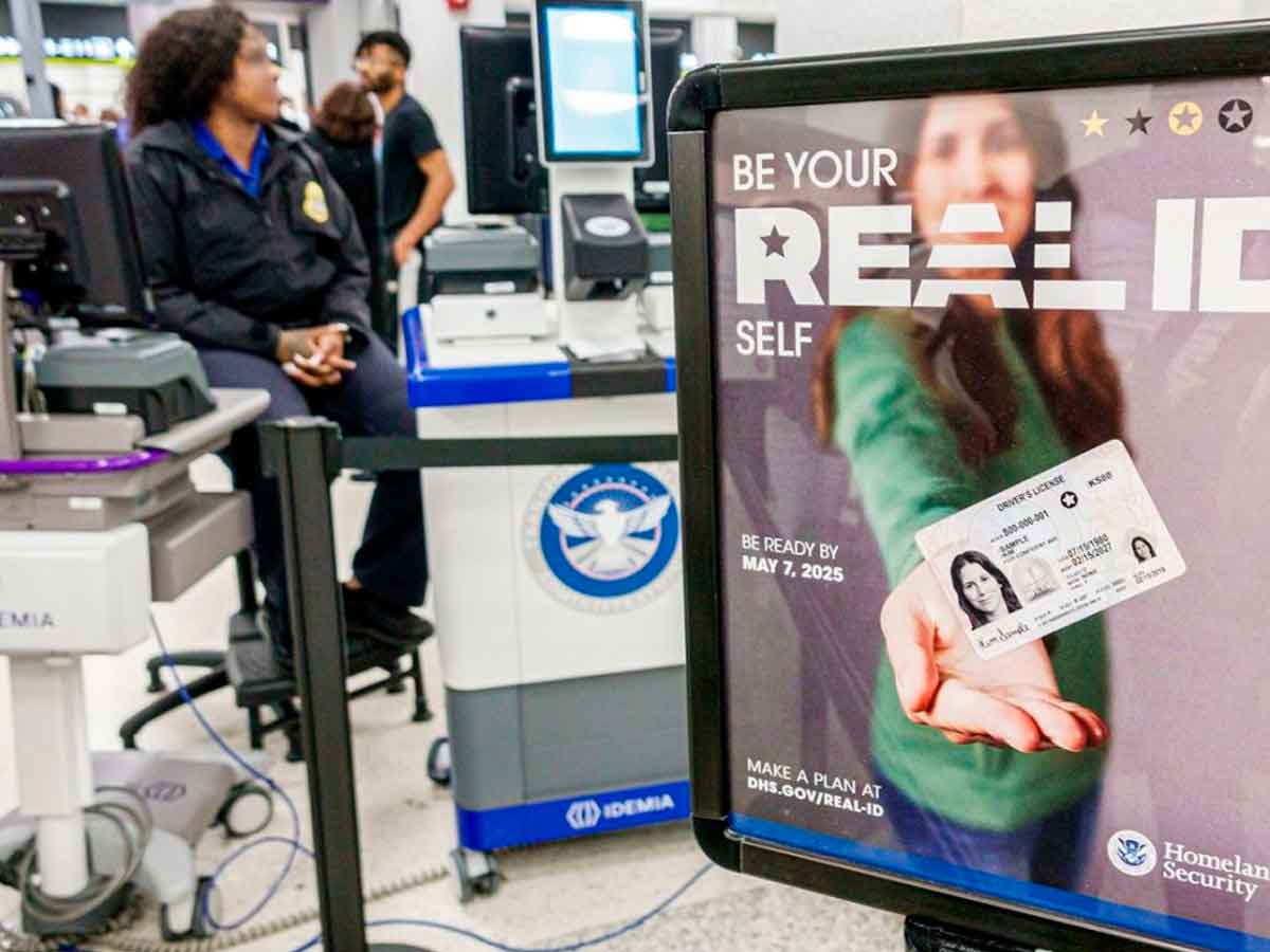 Airport TSA checkpoint with DHS poster urging travelers to obtain a Real ID by May 7 2025; security agent seated in background