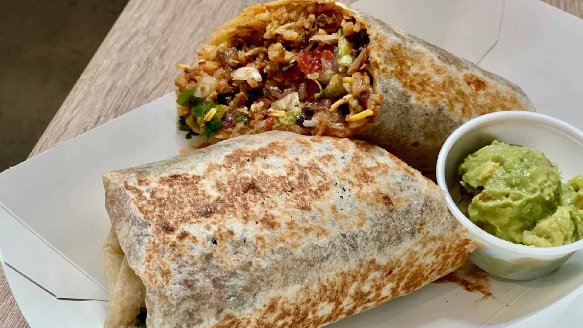Grilled burrito cut in half showing rice, beans and vegetables next to a cup of guacamole in a takeout container