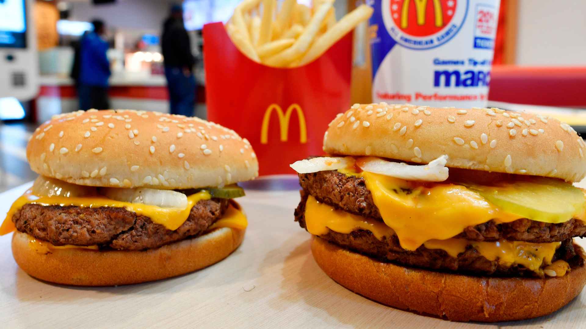 Close-up of two McDonald’s cheeseburgers with fries and branded packaging in a restaurant, highlighting the chain’s new 24/7 service hours.
