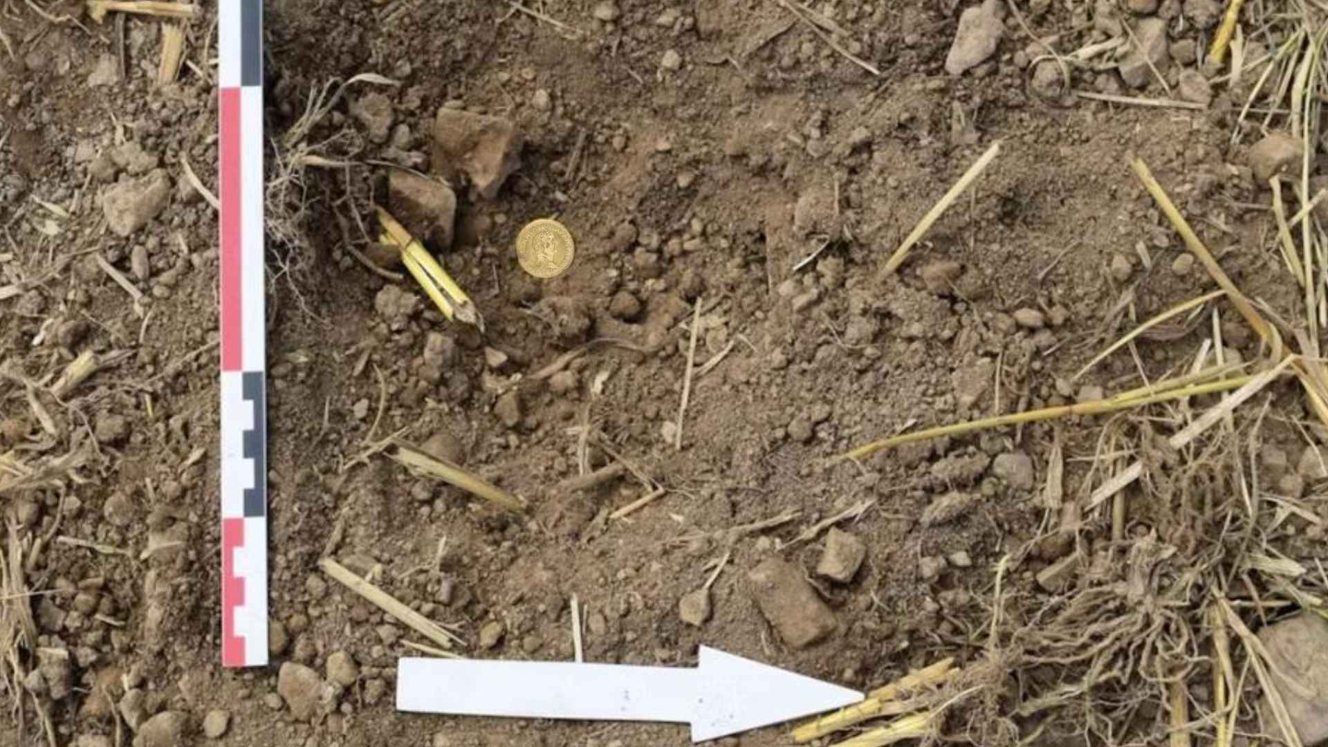 Roman gold coin from 69 A.D. unearthed in Dudley farm soil, beside measurement scale and white arrow marker.