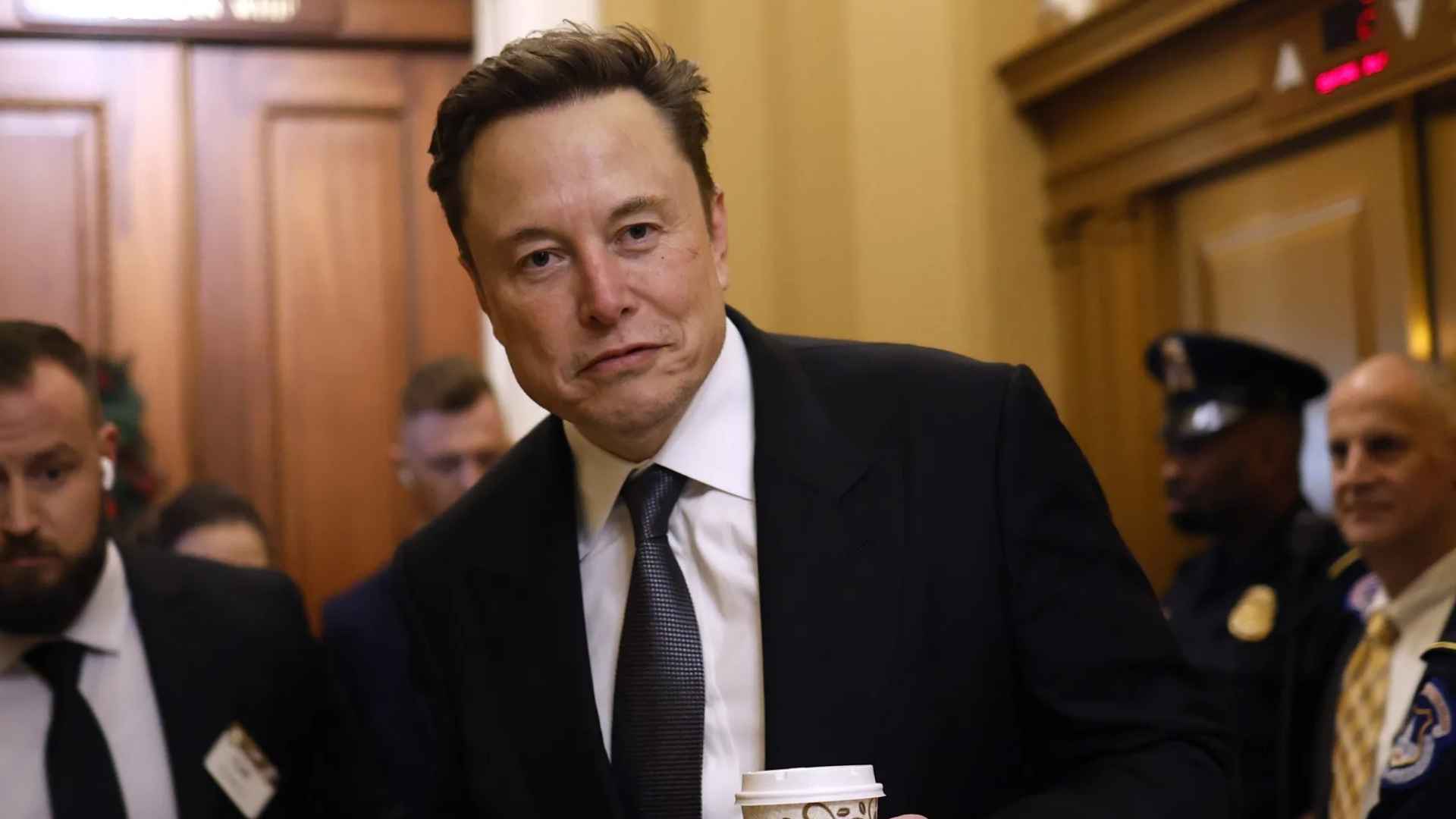 Elon Musk walking through capitol hallway holding coffee with security officers behind him