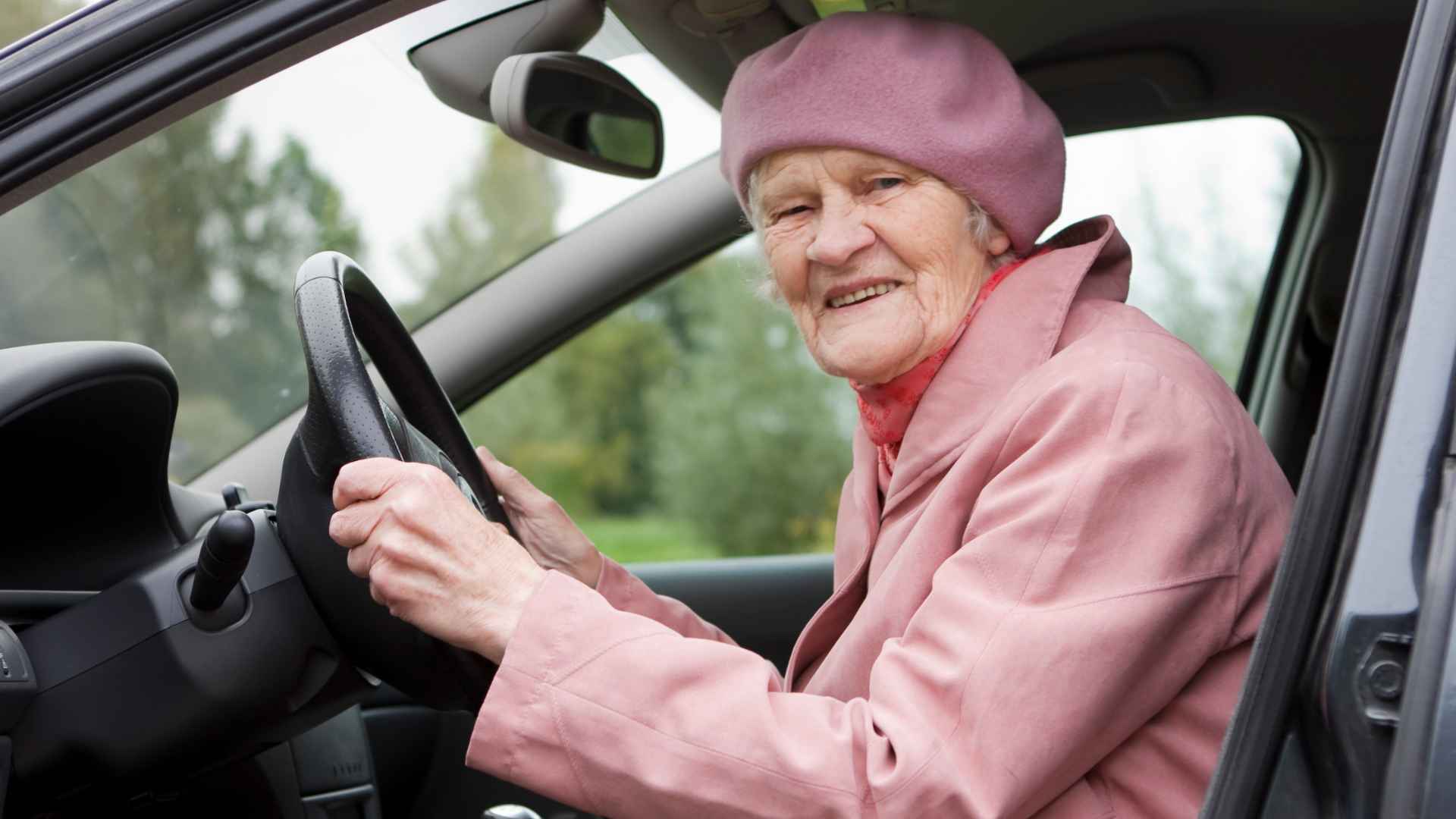 Elderly woman in pink coat grips steering wheel inside car, representing California senior drivers facing DMV renewal rules.