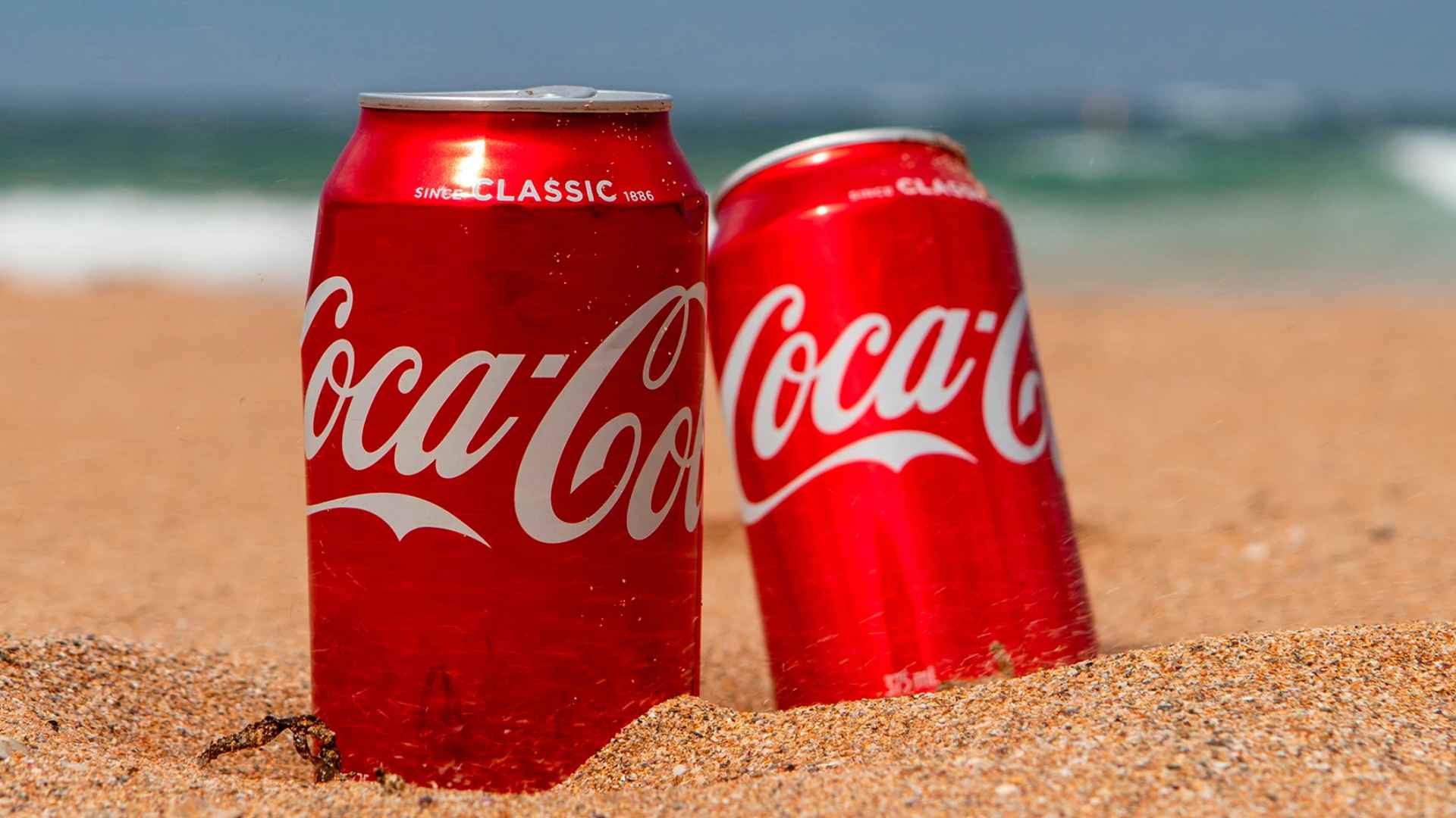 Two red Coca-Cola cans half-buried in sun-lit beach sand with soft-focus ocean waves rolling in behind them.