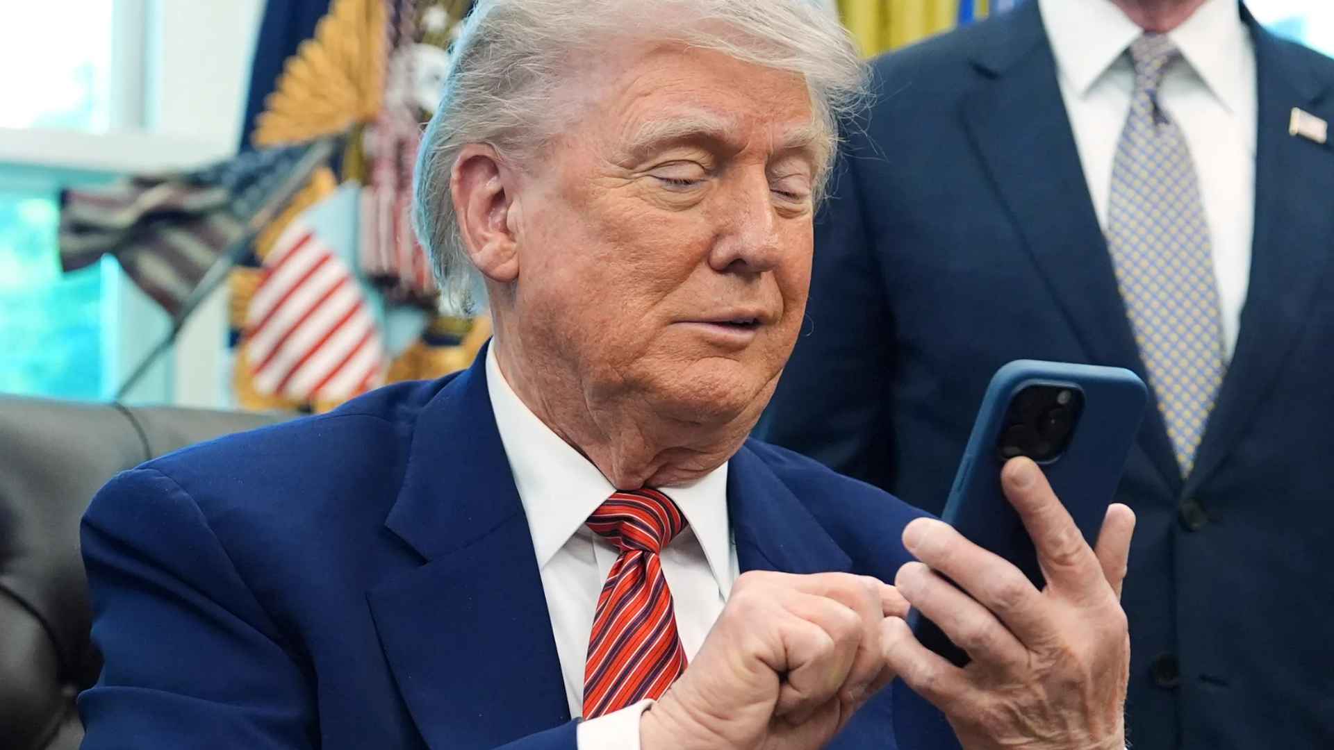 Government official in navy suit checks a smartphone inside the executive office, with U.S. flags blurred in background.