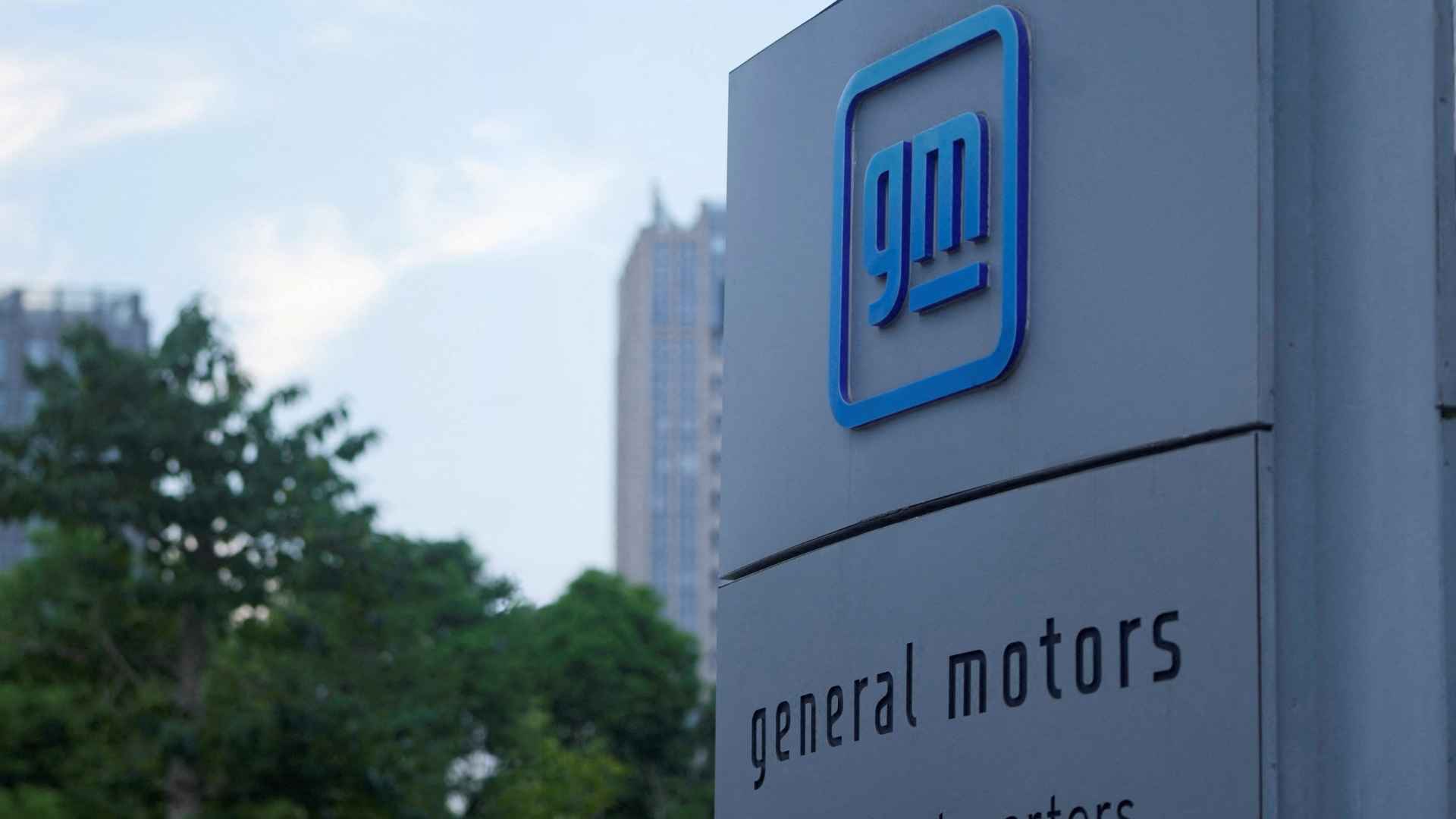 General Motors China headquarters sign with blue GM logo, Chinese flag and city skyline, illustrating automaker’s 600,000-vehicle V8 recall and lawsuit