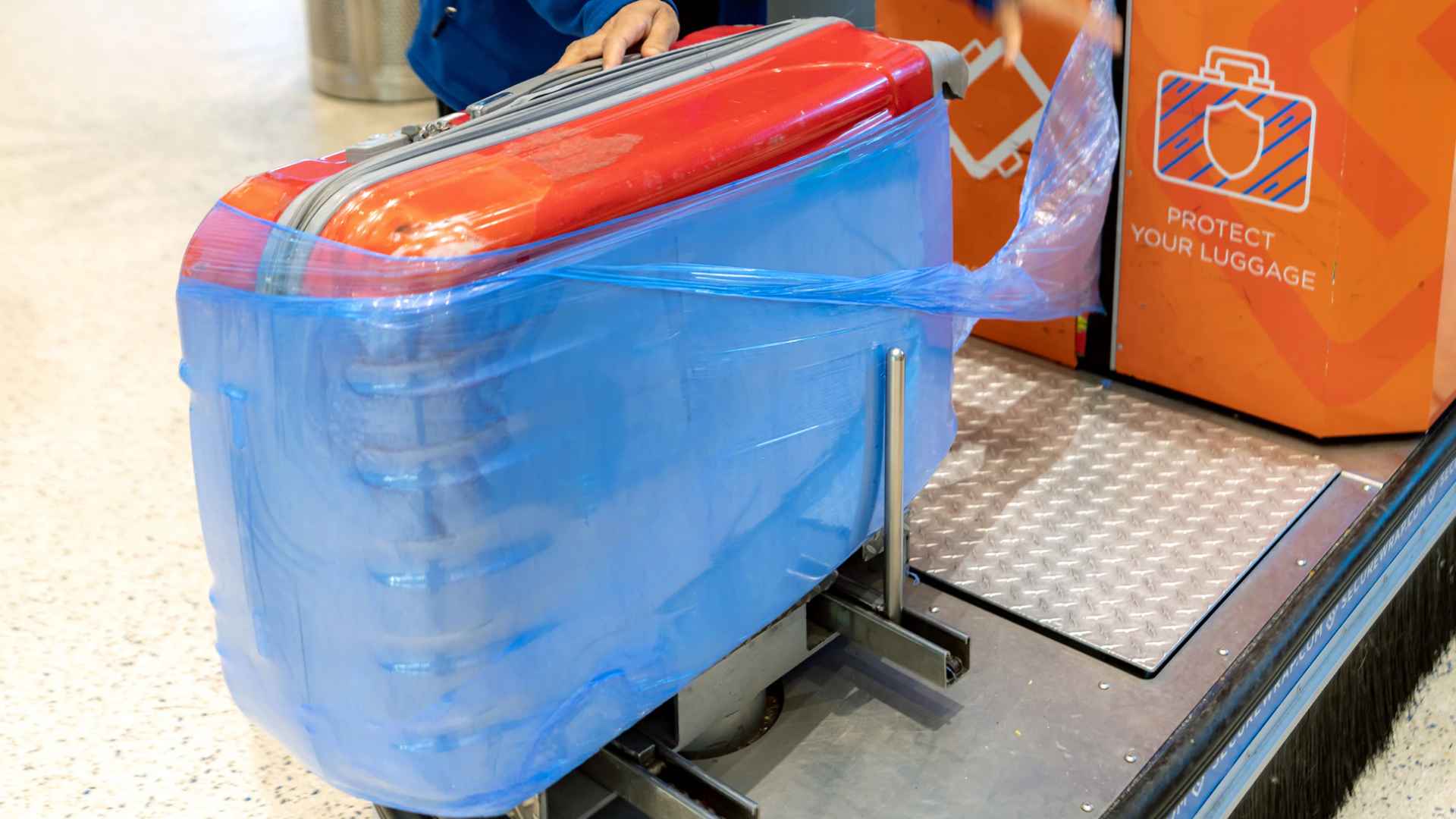 Suitcase wrapped in blue plastic film at an airport check-in station, illustrating TSA advice to skip plastic wrapping