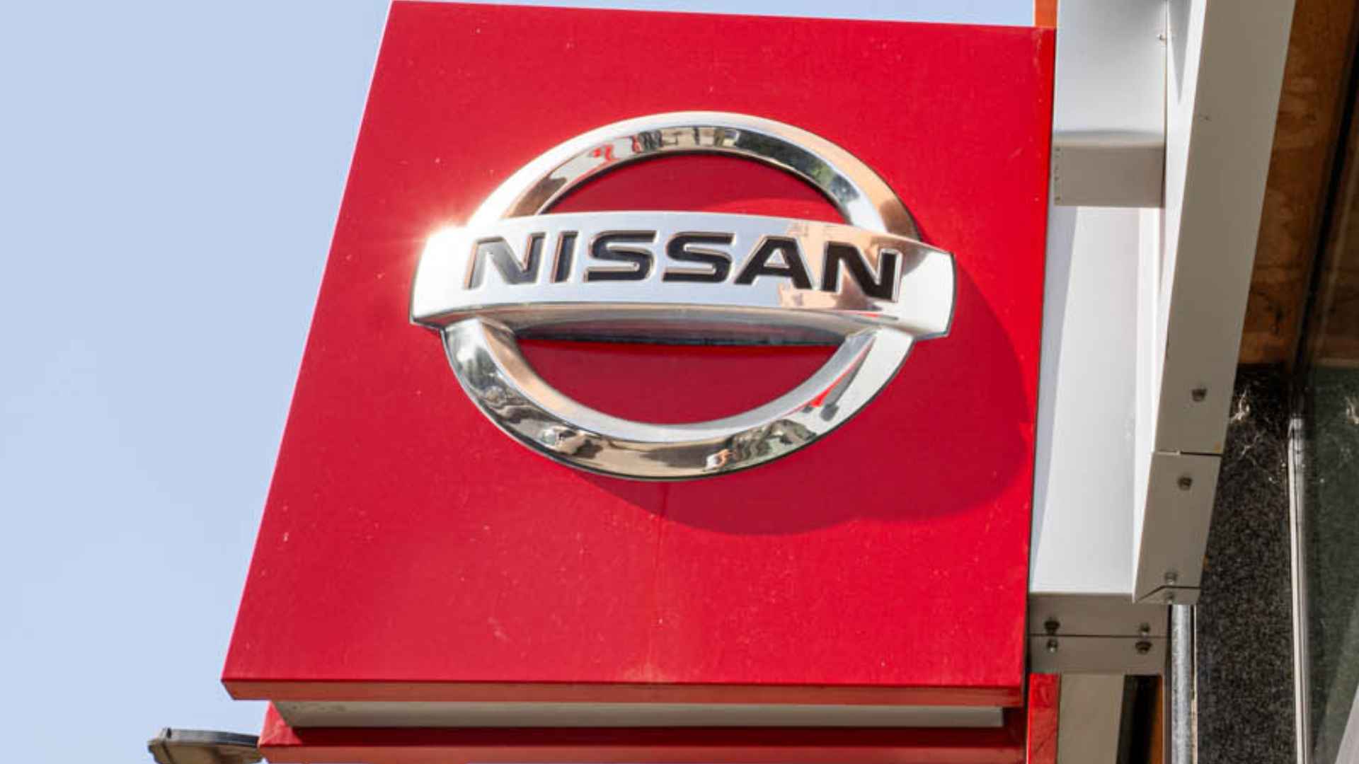 Red Nissan dealership sign reflected in showroom window, illustrating recall of roughly 80,000 2025 Frontier and Kicks vehicles over rear-camera failure.