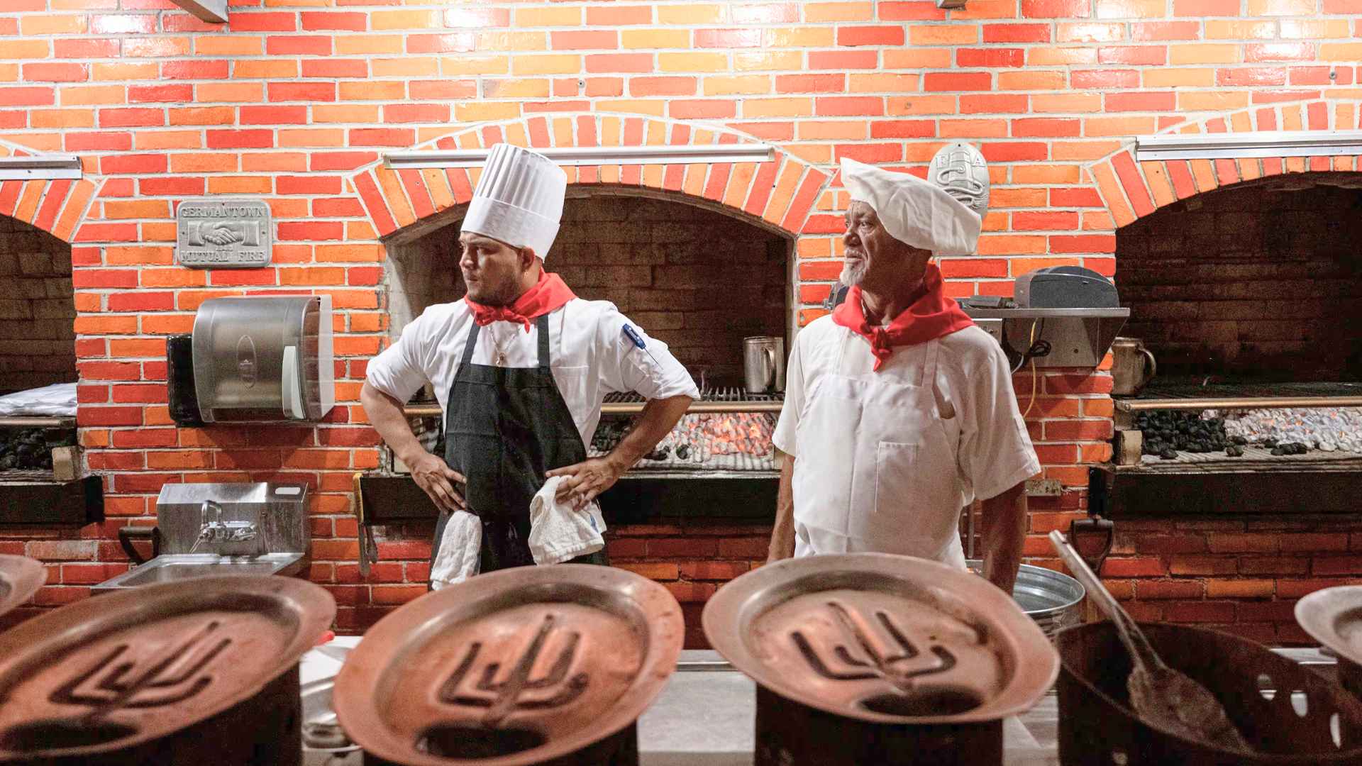 Two chefs in white uniforms stand beside The Pub’s signature open-charcoal fireplace grill inside the historic Pennsauken steakhouse