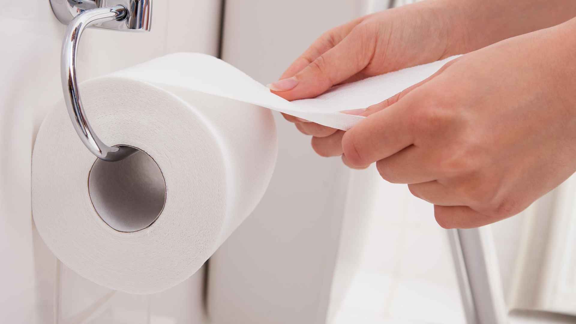 Person unrolling a white toilet-paper roll from a chrome holder in a modern home bathroom.