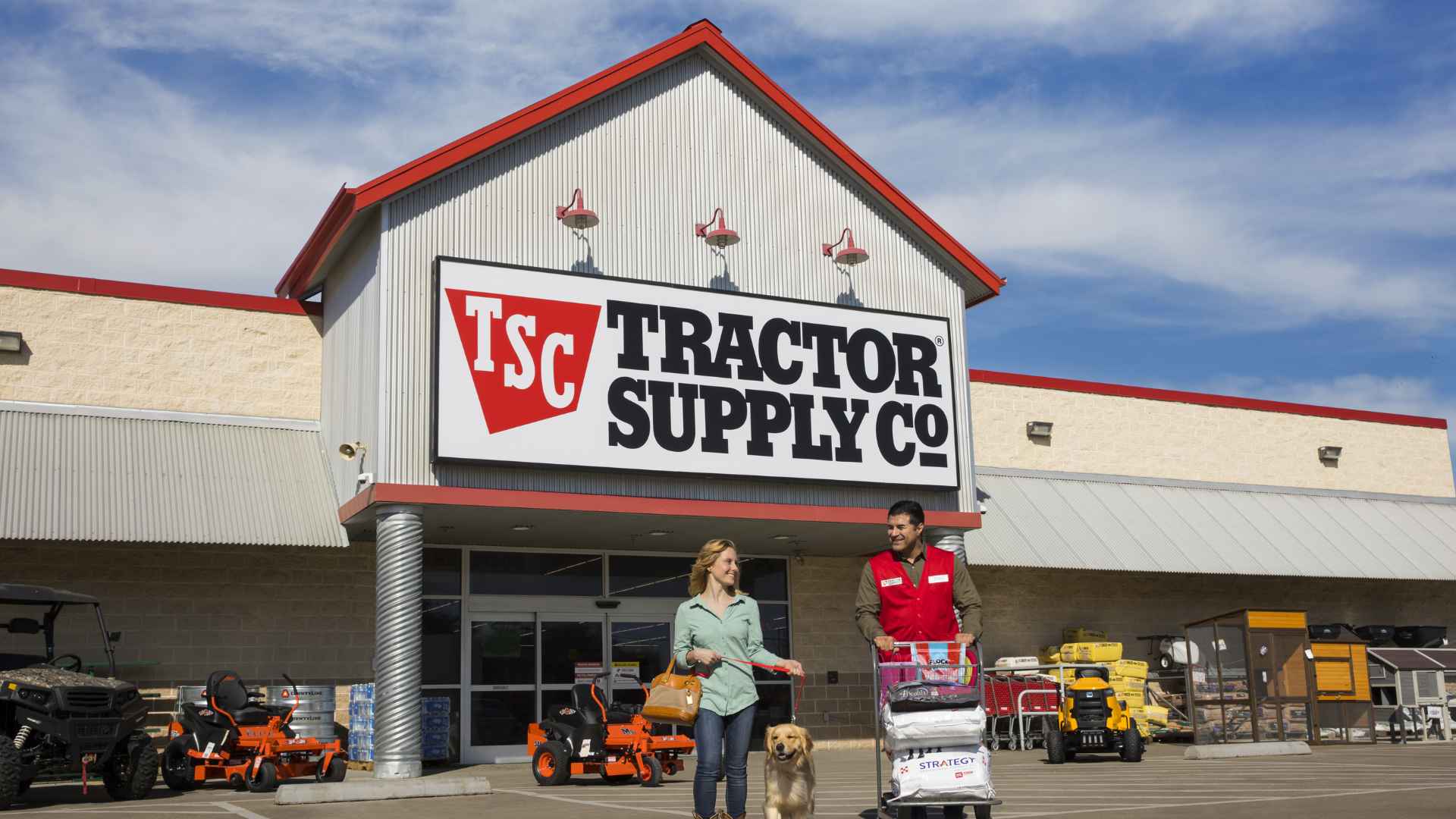 Tractor Supply store exterior with employee helping customer and dog outside, surrounded by outdoor equipment