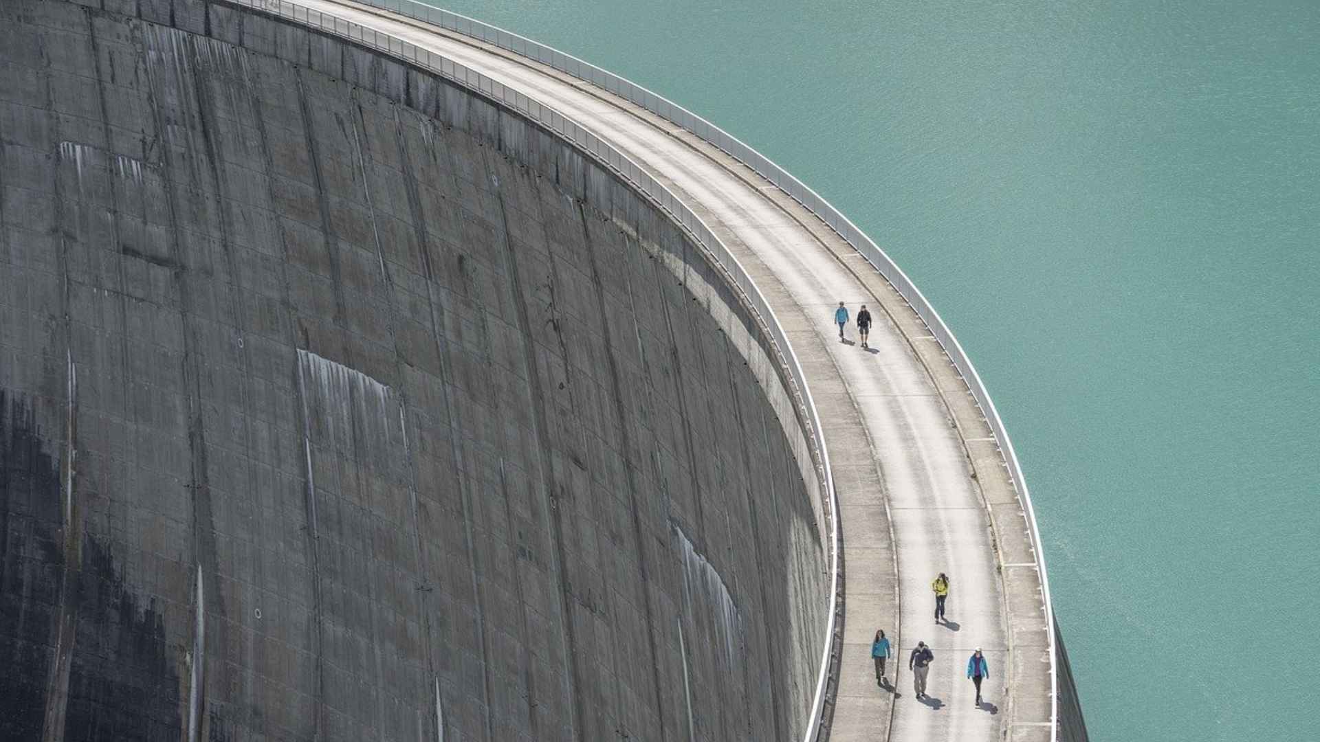 People walking on top of a massive curved hydroelectric dam beside turquoise reservoir water