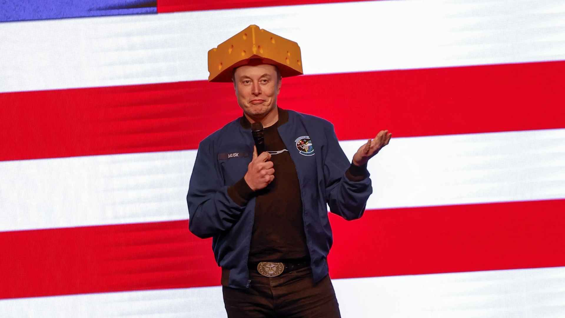 Elon Musk wearing a cheese-shaped hat and shrugging during a speech on Tesla Autopilot issues with NHTSA