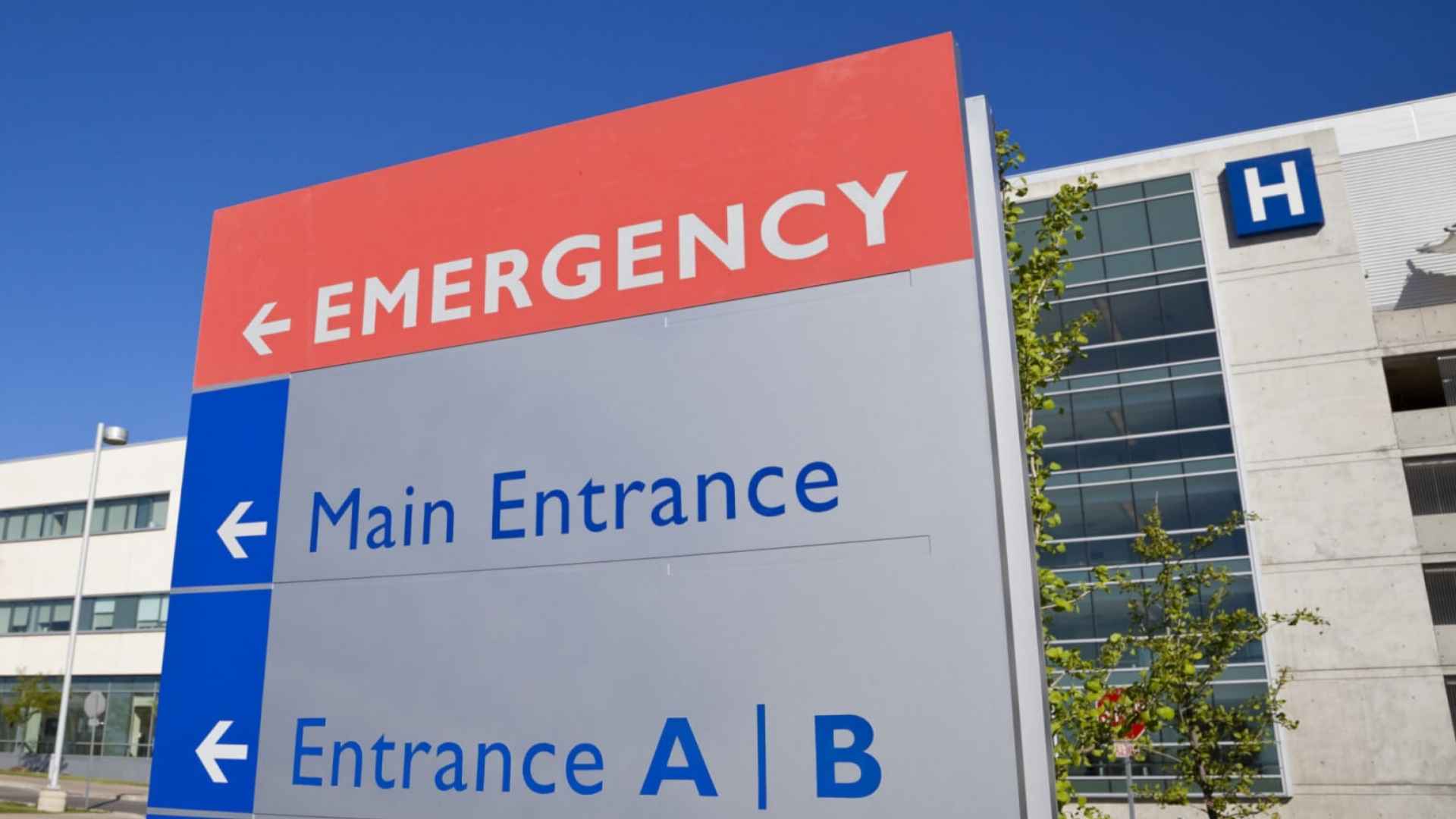 Signpost indicating emergency, main, and A/B entrances outside a modern hospital building