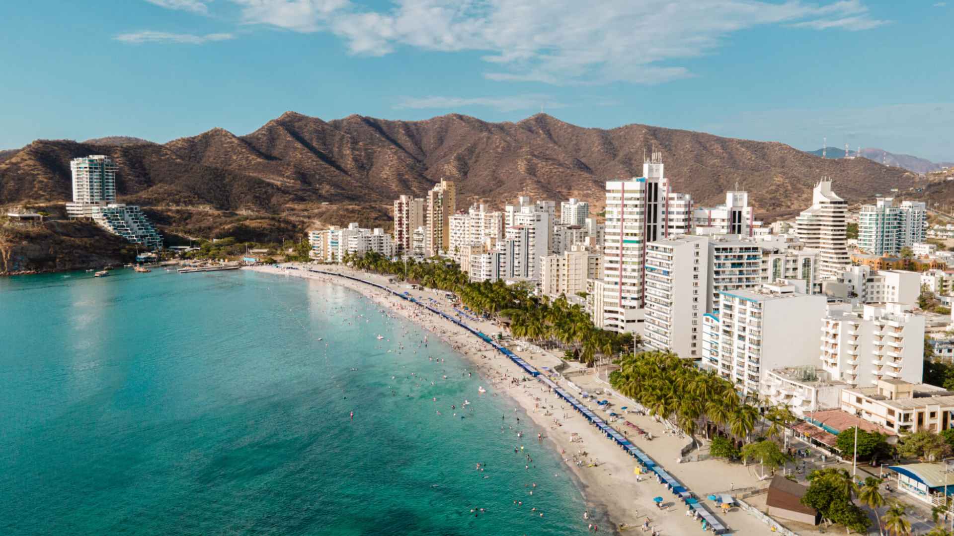 A panoramic view of Santa Marta, Colombia, showcasing the beach, turquoise Caribbean waters, modern buildings, and mountains.