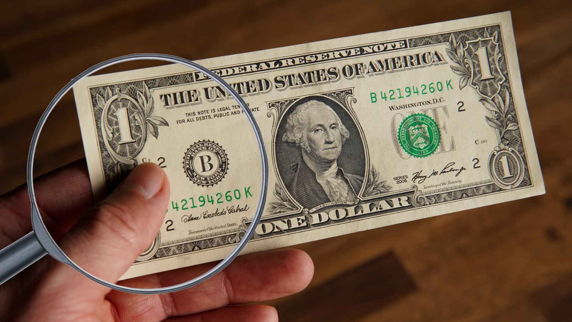 Magnifying glass inspecting a rare 2014 one-dollar bill with duplicate serial numbers, potentially worth $150,000.