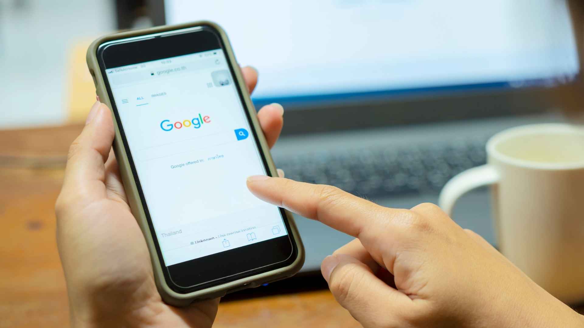 Hand holding smartphone displaying Google homepage with a laptop and coffee mug in the background