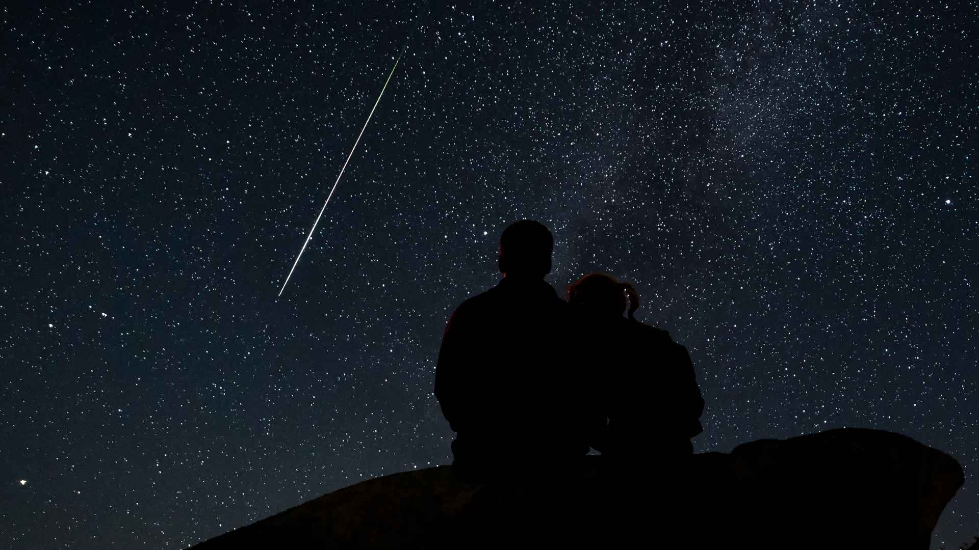Couple watching the Perseid meteor shower in a dark sky with bright shooting stars in August 2025.