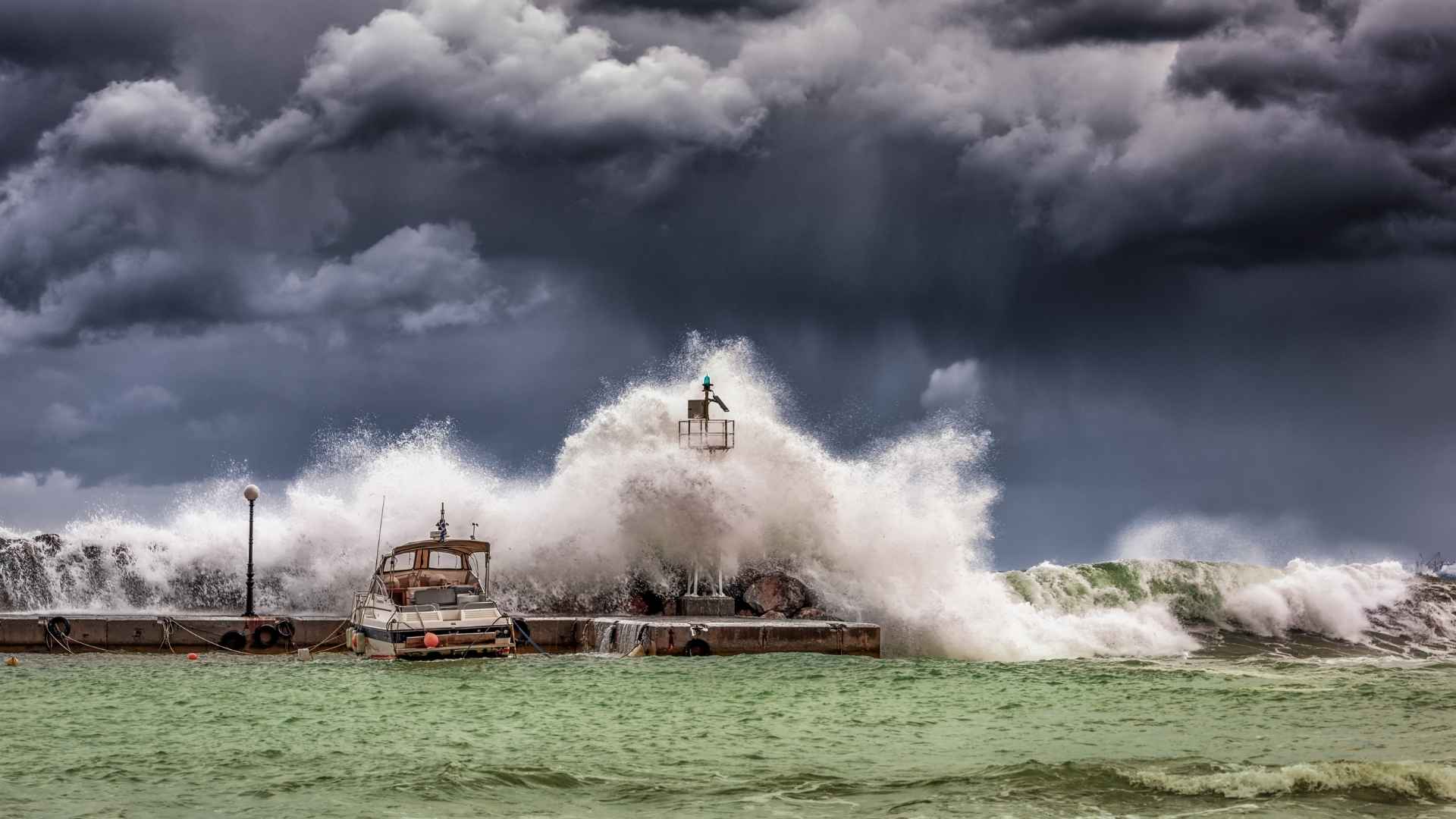 Large waves crash against a pier with a lighthouse during a storm under dark Atlantic clouds in August.