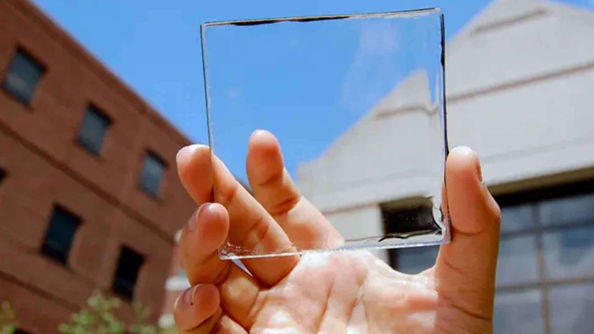 Hand holding a piece of transparent solar glass in front of buildings under a clear blue sky