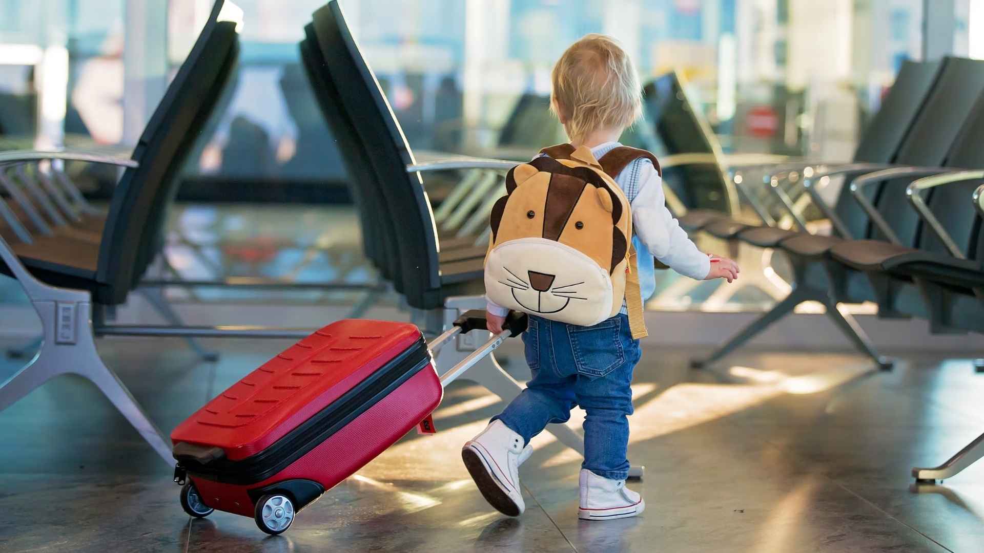 Toddler with animal backpack pulling red suitcase through airport terminal before TSA family security screening