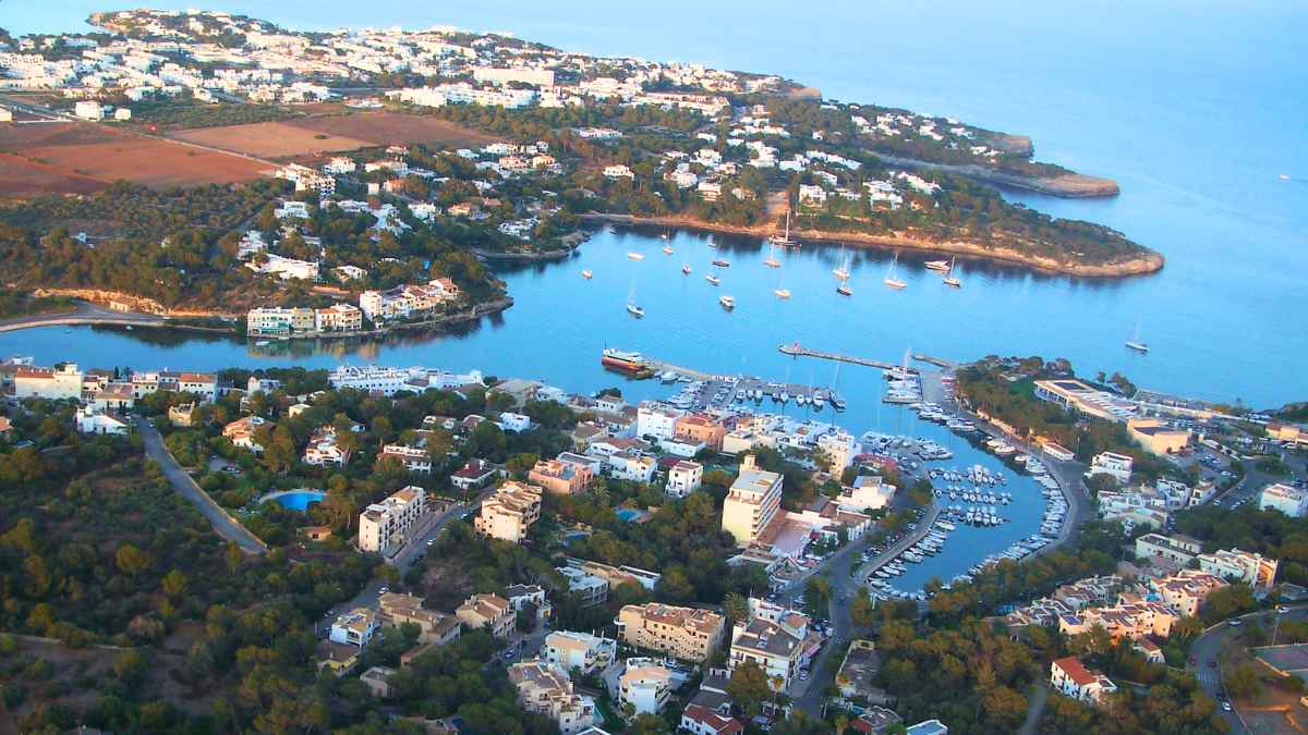 Vista aérea de un pueblo costero en Mallorca con puerto y viviendas rodeadas de naturaleza, donde se ofrecen empleos de vigilante de seguridad