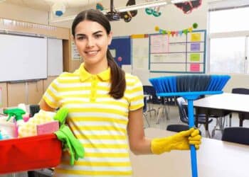 Mujer con uniforme de limpieza sosteniendo una escoba y productos de limpieza en un aula escolar limpia