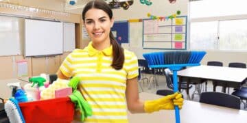 Mujer con uniforme de limpieza sosteniendo una escoba y productos de limpieza en un aula escolar limpia