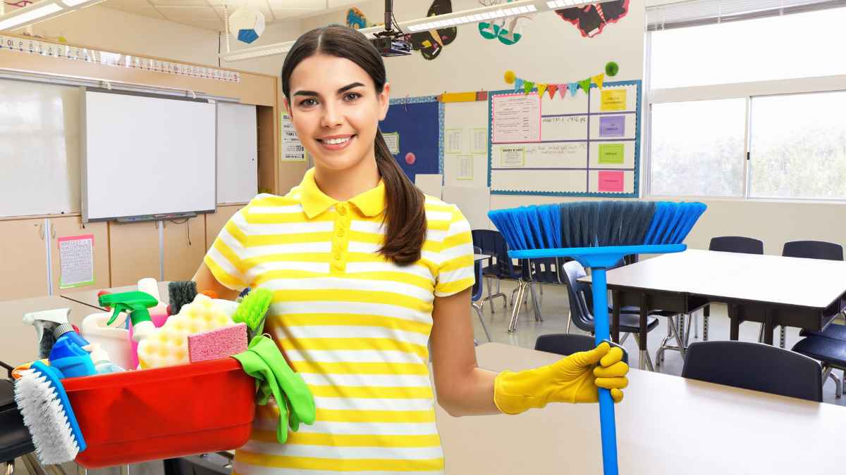 Mujer con uniforme de limpieza sosteniendo una escoba y productos de limpieza en un aula escolar limpia