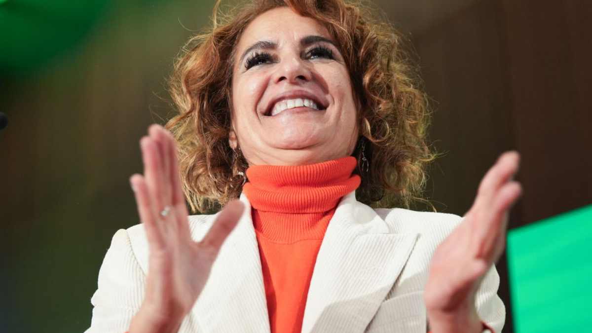 María Jesús Montero con chaqueta blanca y jersey naranja aplaudiendo en un acto sobre las devoluciones del IRPF a mutualistas.