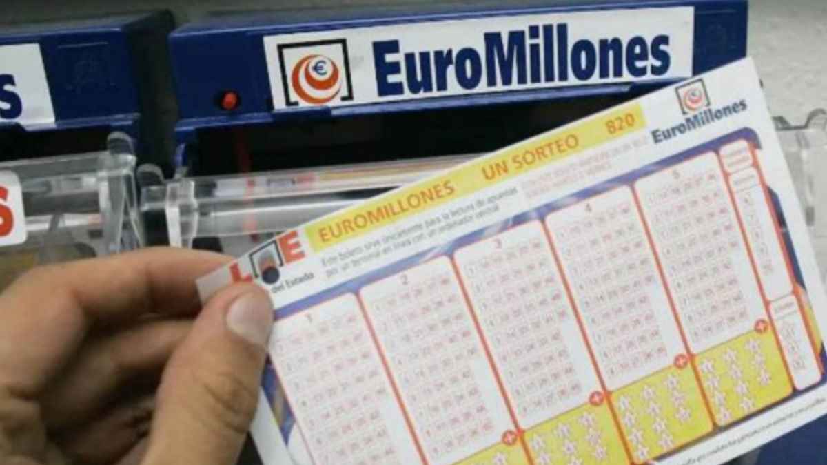 Mano sosteniendo un boleto de Euromillones frente al mostrador de Loterías y Apuestas del Estado
