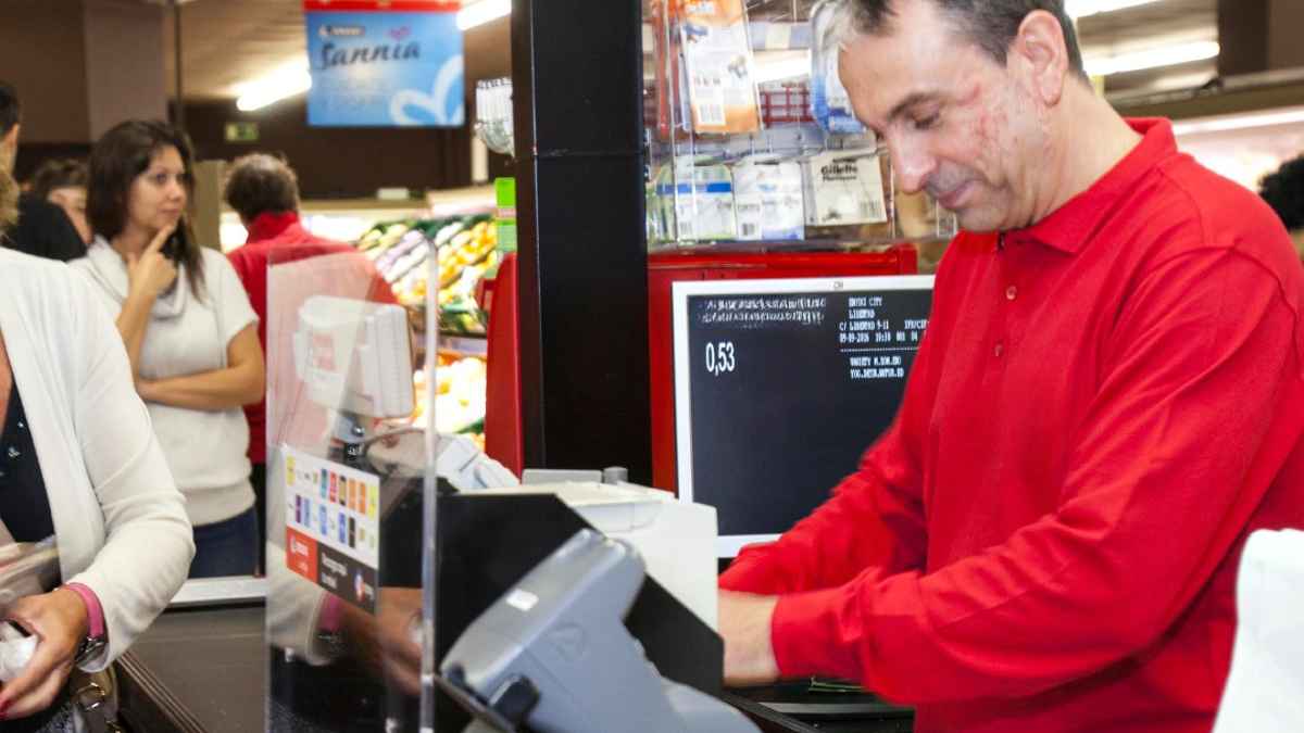 Cajero con polo rojo en la caja registradora intentando cobrar a clientes, caso de despido para cobrar el paro