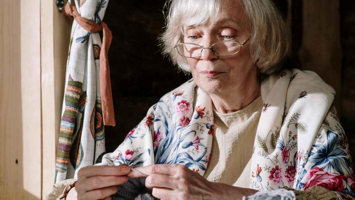 Trabajadora de 65 años con gafas y chal floral tejiendo junto a la ventana, símbolo del despido por negarse a jubilarse
