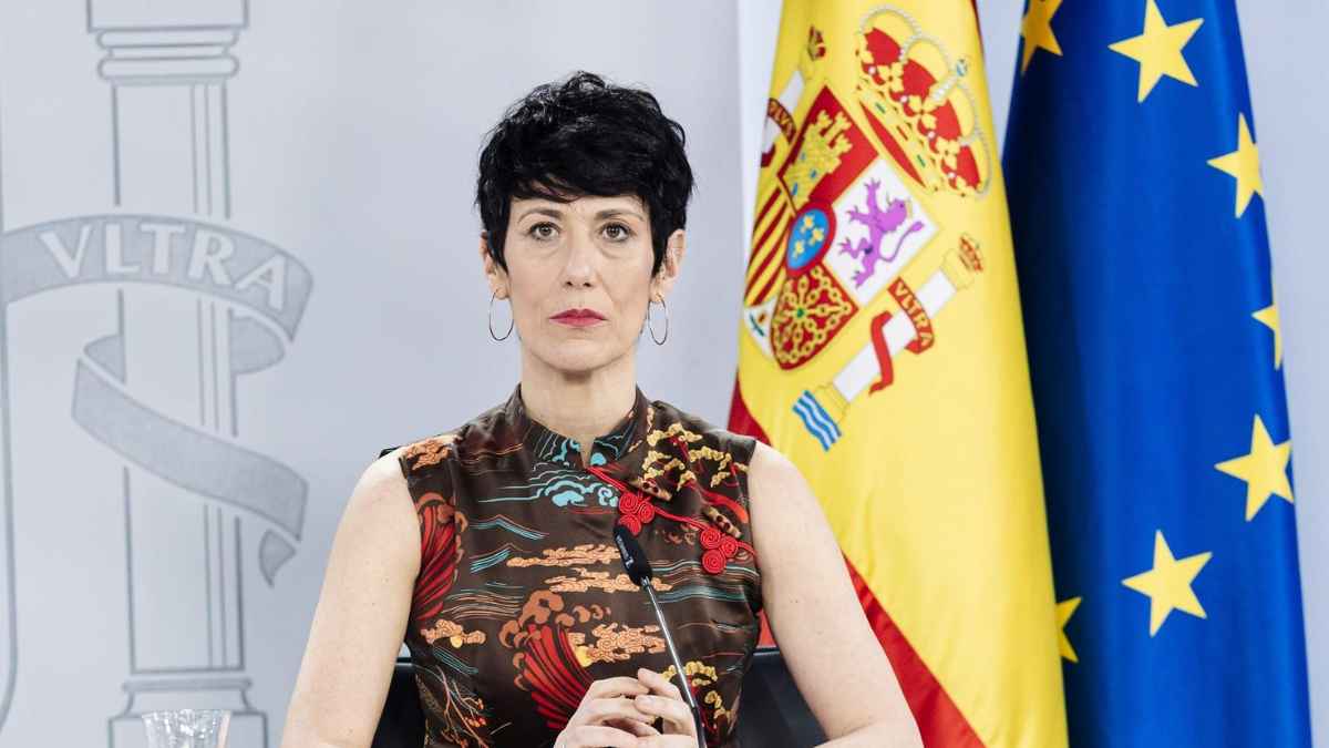 Representante del Gobierno de España, flanqueada por las banderas nacional y europea, anuncia coeficientes reductores del 30 % en pensiones por jubilación anticipada involuntaria.