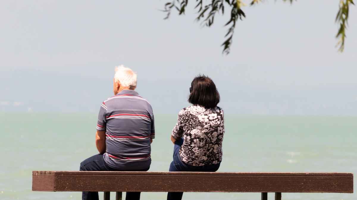 Pareja mayor sin jubilación en banco frente al mar.