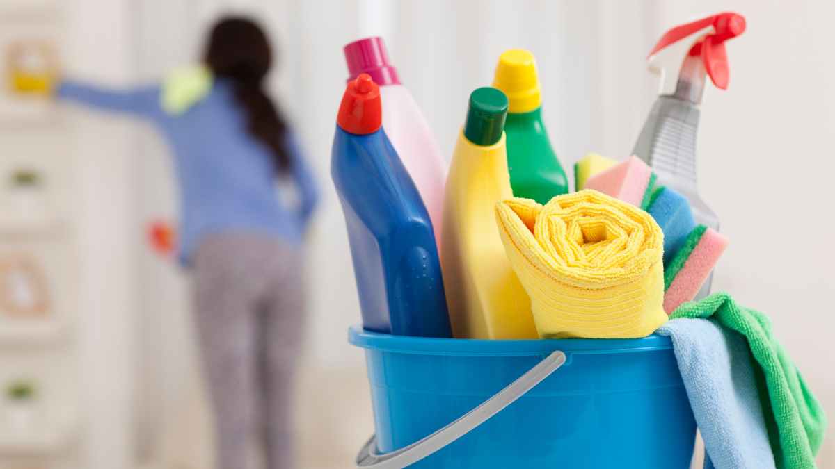 Productos de limpieza en un cubo azul con una empleada del hogar difuminada al fondo durante sus tareas.