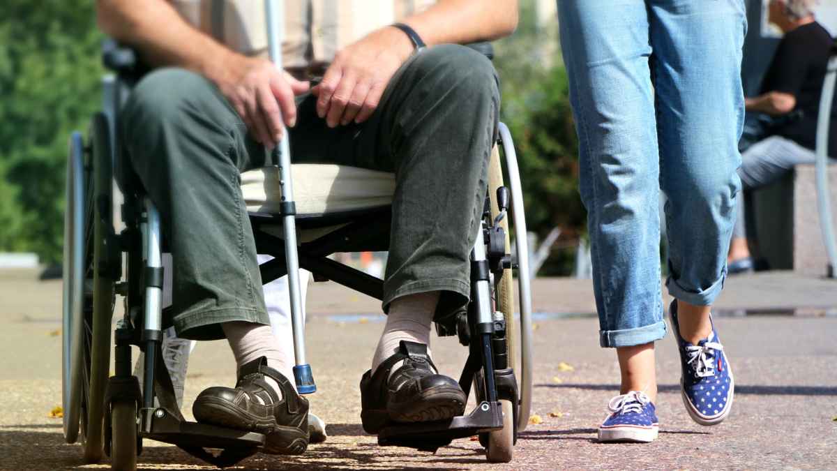 Hombre en silla de ruedas con muleta, acompañado mientras se desplaza; símbolo de beneficiario de incapacidad permanente absoluta y empleo incompatible con Seguridad Social.