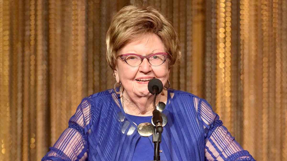 Joan Payden, millonaria de 92 años con vestido azul hablando en público frente a un micrófono
