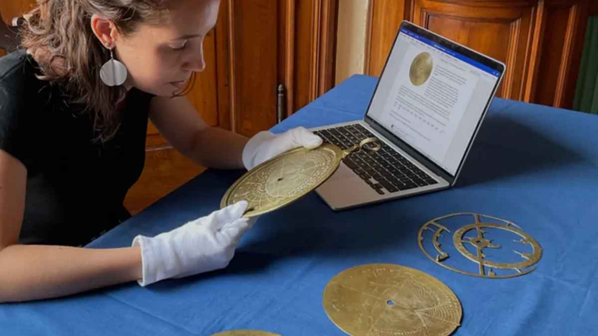 Historiadora sosteniendo un astrolabio antiguo de más de 1.000 años sobre una mesa azul, junto a piezas del instrumento y un portátil que muestra información.