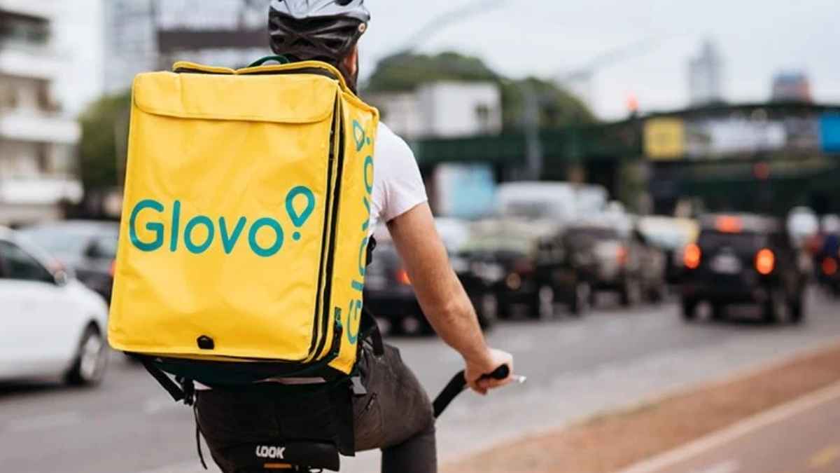 Trabajador de glovo en bicicleta con mochila amarilla en una avenida urbana
