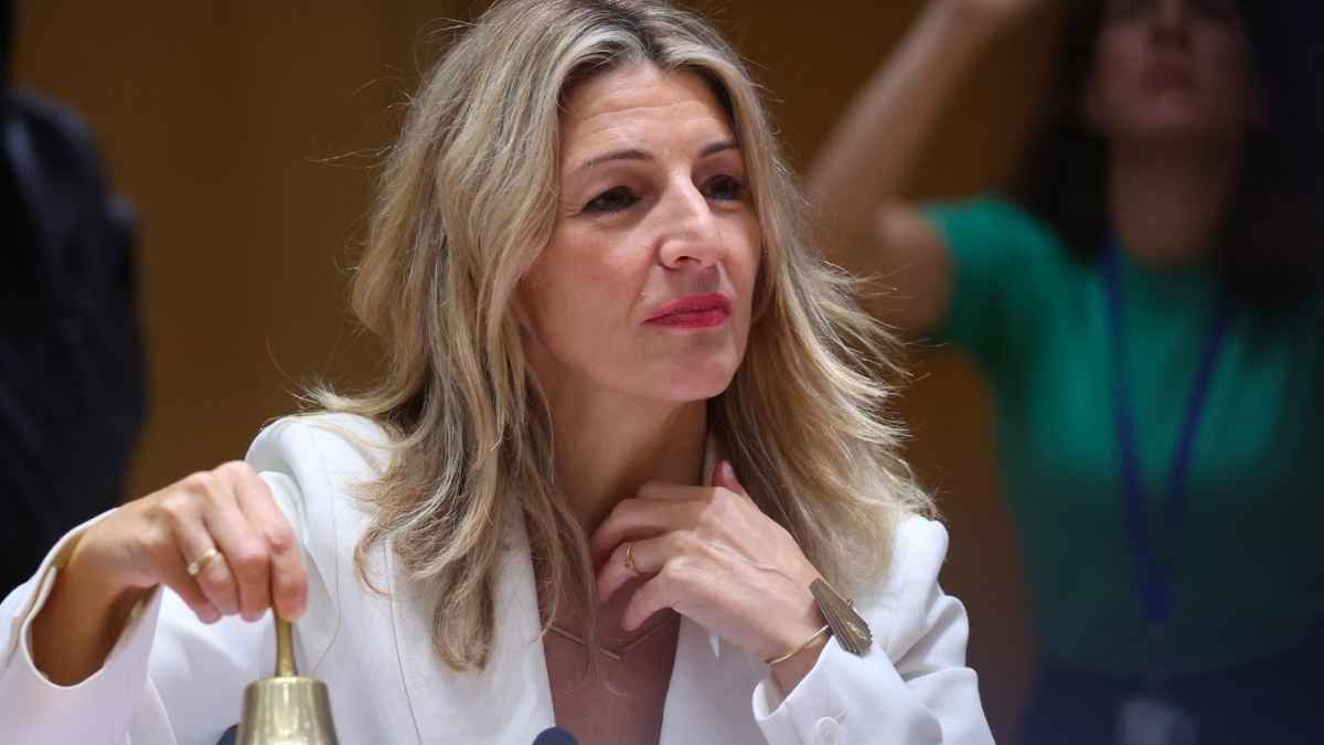 Ministra Yolanda Díaz participando en una reunión, mostrando un gesto reflexivo mientras sujeta una campana.
