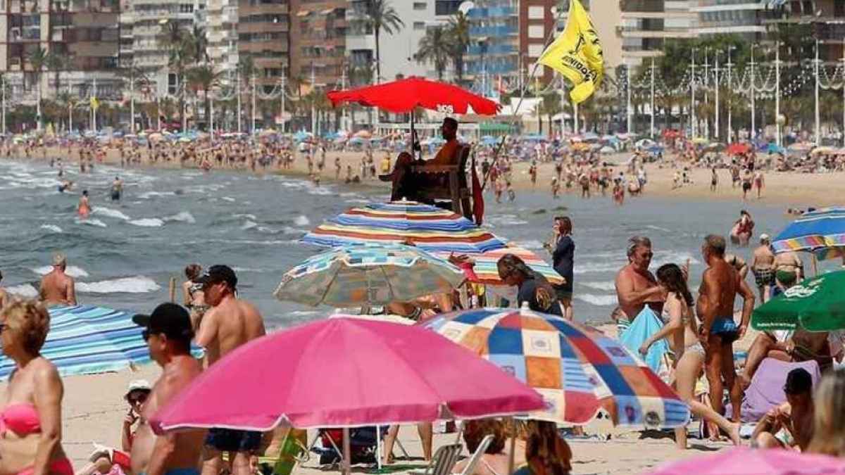 Turistas con sombrillas y hamacas reservando sitio en la concurrida playa de Benidorm durante el verano.