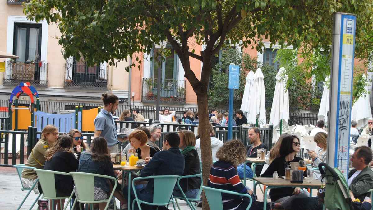 Camarero atiende varias mesas en una terraza al aire libre, junto a un parque infantil, con gran afluencia en hora punta.