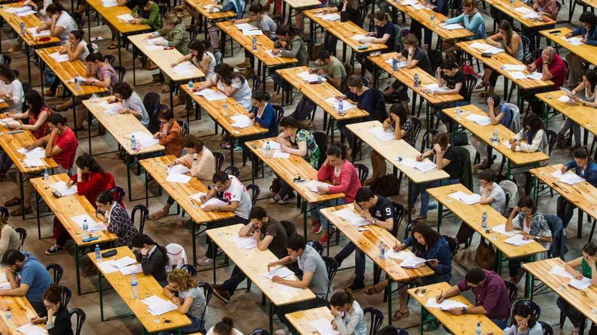 Personas realizando examen de oposiciones en gran sala con cientos de aspirantes