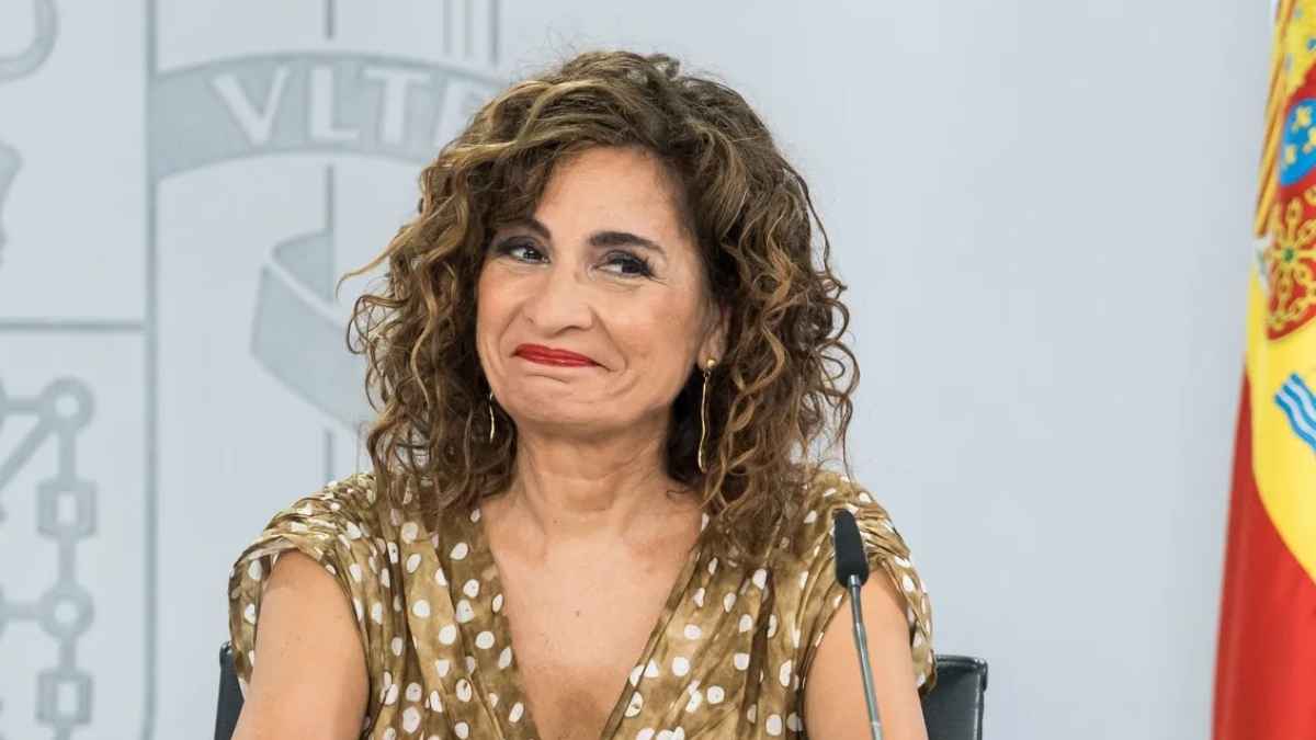 Mujer en rueda de prensa con gesto sonriente ante un fondo institucional y bandera de España