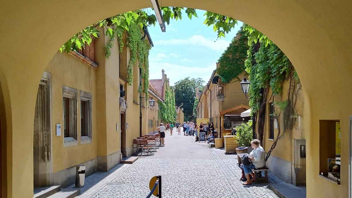 Calle interior del barrio histórico Fuggerei en Augsburgo con casas amarillas, fachadas con hiedra y visitantes paseando