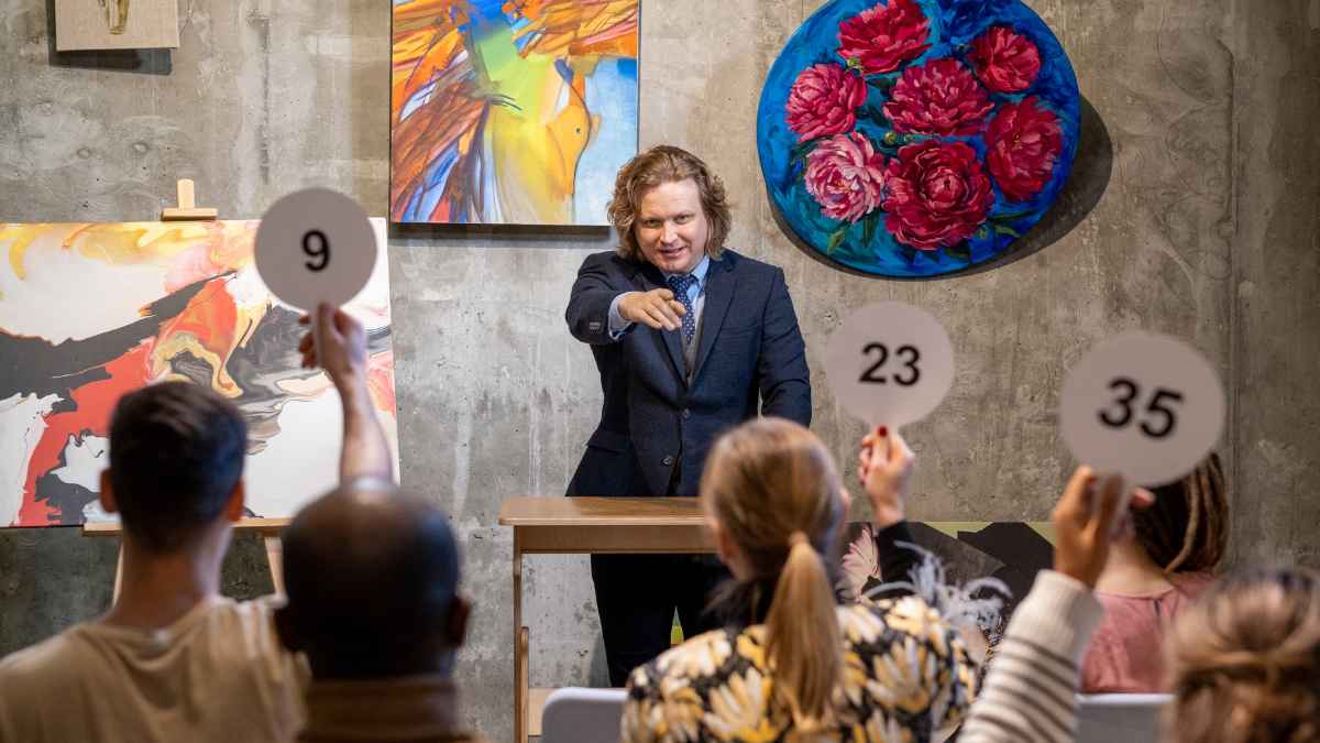 Subasta de arte con un subastador señalando y varios asistentes levantando paletas numeradas