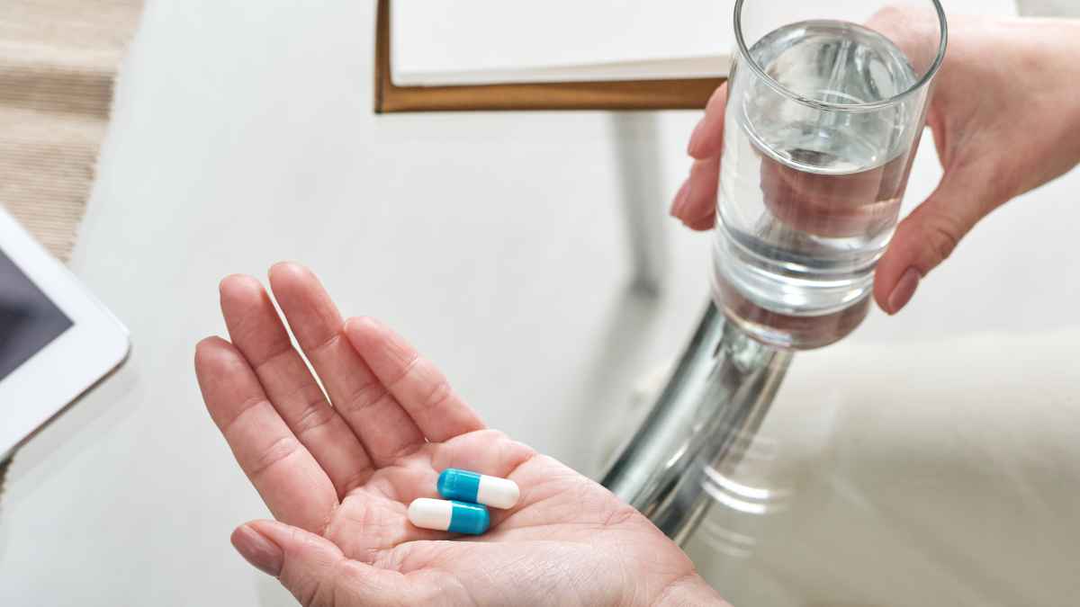 Mano sosteniendo dos cápsulas junto a un vaso de agua listo para tomar medicación