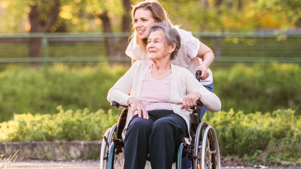 Mujer joven empujando la silla de ruedas de una persona mayor en un entorno al aire libre mientras la cuida.