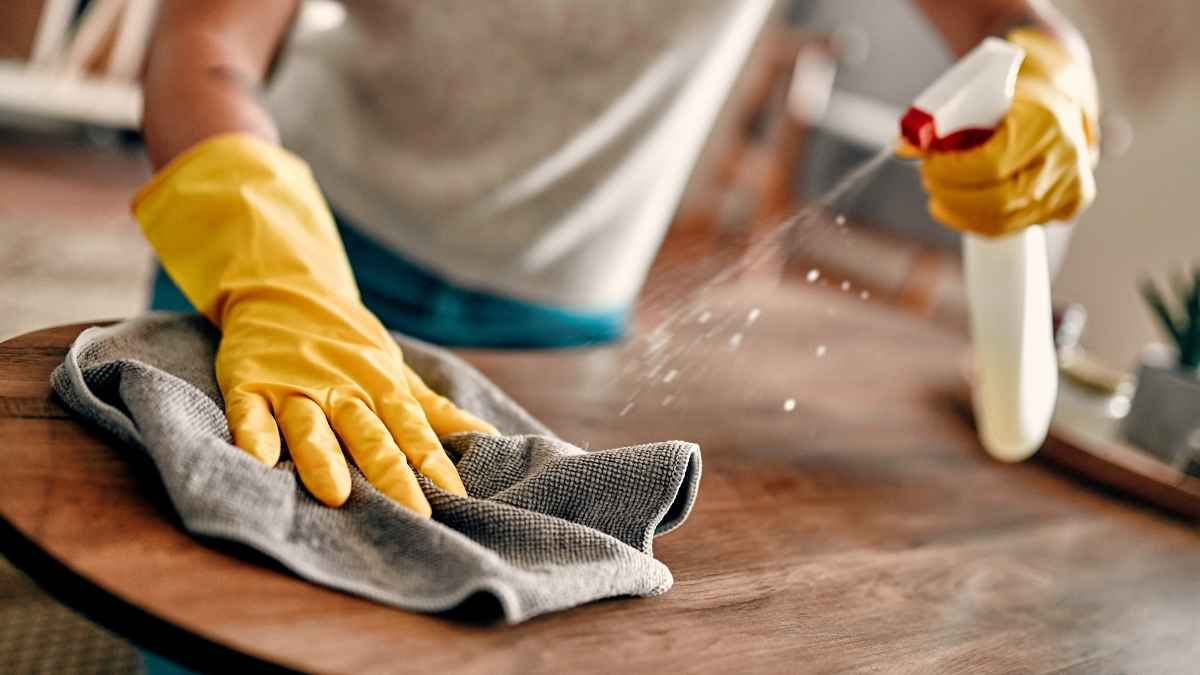 Persona limpiando una mesa con guantes amarillos y spray durante una jornada de trabajo en el hogar