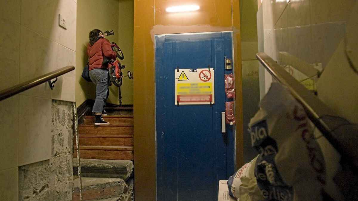 Persona subiendo una bicicleta por las escaleras de un edificio sin accesibilidad junto a un ascensor antiguo.
